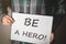 Young man in plaid shirt holding a placard with text: BE A HERO!