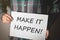 Young man in plaid shirt holding a placard with text: MAKE IT HAPPEN!