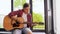 Young man playing guitar sitting on windowsill