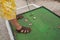 Young man playing minigolf on old, worn hotel resort green field, closeup to his feet in sandals, metal club and ball