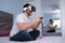 Young man playing video games with virtual reality headset and gamepad sitting on bed, reflection on wardrobe mirror