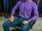 A young man in a purple sweater and jeans sits in a brown leather armchair against the background of a wardrobe with clothes