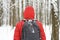 Young man in a red jacket with a hood and backpack in a snowy winter forest or Park, rear view from the back.