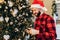 Young man in Santa Claus hat, uses a mobile phone, dials sms, has merry Christmas, a Christmas tree at home