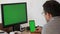 A young man in a shirt and glasses sits in front of a green chroma key monitor screen and clicks something on a