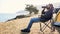 Young man sits on chair at campsite on lake shore and drinking from thermos