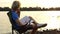 Young Man Sits, Dreams, Smiles, Keeps a Notepad, on a Riverbank