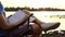 Young man sits, dreams, writes something into a notepad, on a riverbank