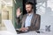 A young man sits in the office at a desk in a headset and works on a laptop, talking and greeting to the camera online