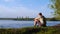 Young man sits on shore of river on background of city. Stock footage. Man enjoys views of nature sitting by river
