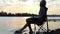 Young Man Sits And Smiles And Moves His Barefoot in Water on a Riverbank