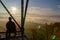 Young man sitting from back on watchtower construction looking to autumn foliage trees with misty fog and hill at sunrise, Czech