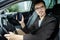 Young man is sitting at his car while looking at the camera. His left hand is on the steering wheel. He is holding the phone in