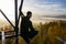 Young man sitting from side on watchtower construction looking to autumn foliage trees with misty fog and hill at sunrise, Czech