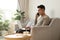 Young man sitting on sofa at home with laptop talking with friend on smartphone