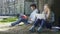 Young man sitting under tree with book, looking at girl with laptop, indecisive