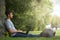 Young man sitting under a tree with his legs stretched and his computer on them. Technological concept.