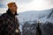 Young man standing on the edge of a mountain cliff, using an action camera to take a photo of snowy mountains.