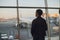 Young man is standing near window at the airport and watching plane before departure. Focus on his back