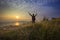 Young man standing and rising hand as victory on grass hill looking to sun above sea horizontal with dramatic colorful sky