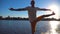 Young man standing at yoga pose on wooden jetty at lake. Athlete balancing on one leg at nature. Sporty guy doing