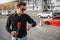 Young man stands outside on street and hold red rose. He looks at watches. Guy is waiting. There is road with cars