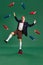 Young man in stylish clothes and glasses balancing on books over flying books against green studio background