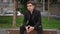 A young man in a suit with a serious face sits on a bench in a city park. The man is looking forward to the meeting with