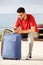 Young man with suitcase sitting outside listening to music with cellphone and headphones