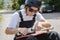 A young man in sunglasses and overalls with a helmet on his head changes his wheels on his longboard under the open sky