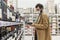 A young man in a supermarket chooses alcohol in the wine section. A brunette in a beige coat with a beard wearing a medical mask