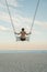 Young man on swing high in the sky. Clouds background. Guy swinging back view. Soft light of sunset. Back view