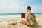 Young man in tank top, shorts, snapback cap at beach with laptop & backpack. Freelancer, hipster blogger, writer enjoying sea view