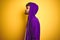 Young man with tattoo wearing purple sweatshirt with hood over isolated yellow background looking to side, relax profile pose with