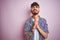 Young man with tattoo wearing striped shirt standing over isolated pink background begging and praying with hands together with