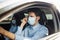 Young man taxi driver adjusts sterile medical mask in the car. A boy behind the car steering wheel safely driving for business