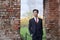 A young man, a teenager, in a classic suit. Pondering is standing in front of the old wall of red brick, putting his hands in his