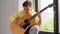 Young man tuning guitar sitting on windowsill