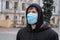 Young man on urban street in protective mask on face. Man in black clothes standing against the background of urban buildings,