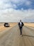 Young man walks on highway, his car at roadside parked in desert. White SUV.