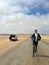 Young man walks on highway, his car at roadside parked in desert. White SUV.