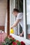 Young man watering blooming flowers looking out of window of country house