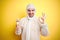 Young man wearing painter equipment and holding painting brush over isolated yellow background doing ok sign with fingers,