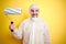 Young man wearing painter equipment and holding painting roller over isolated yellow background with a happy face standing and