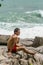 A young man wearing red swimming shorts sitting on the rocks and looking to ocean. Summer resting on the beach.