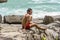 A young man wearing red swimming shorts sitting on the rocks and looking to ocean. Summer resting on the beach.