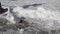 Young man wearing warm trousers and black sneakers is playing with snow by kicking snowdrift in city park on sunny winter