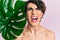 Young man wearing woman make up holding green plant leaf close to beautiful face angry and mad screaming frustrated and furious,