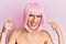 Young man wearing woman make up wearing pink wig looking confident with smile on face, pointing oneself with fingers proud and