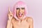 Young man wearing woman make up wearing pink wig pointing finger up with successful idea